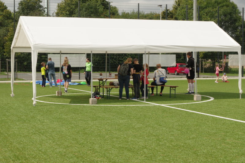 Ein Zelt auf einem Rasenplatz. Darunter Menschen die miteinander sprechen. Im Hintergrund Kinder und Erwachsene die Bewegungsspiele spielen.