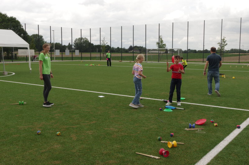 Kinder die auf einem Sportplatz jonglieren. Eine Frau schaut zu.