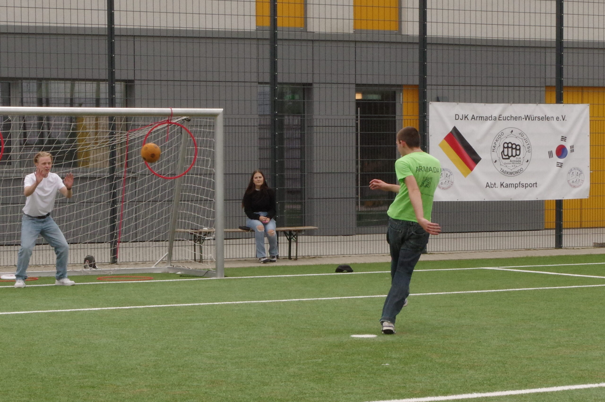 Ein junger Mann schießt mit einem Fusball auf ein Tor. Ein anderer junger Mann steht im Tor und versucht den Ball zu fangen. Im Hintergrund sitzt eine junge Frau.