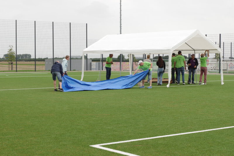 Mehrere Menschen mit grünen T-Shirts bauen auf einem Fußballfeld ein großes Zelt ab.