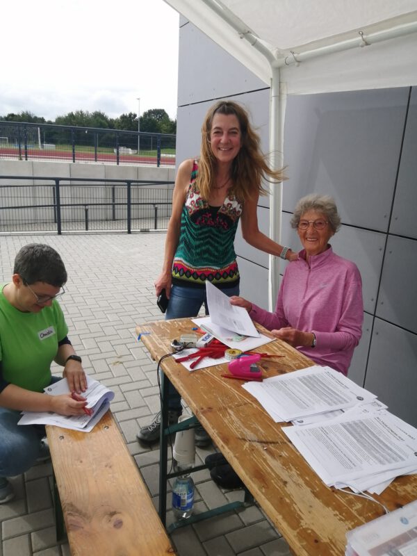 Eine öltere Frau sitzt draußen an einem tisch und hält einen Zettel in der Hand. Neben ihr steht eine Frau die ihr die Hand auf die Schulter legt. links im Bild hockt eine Frau in einem grünen T-Shirt, die etwas auf einen Zettel schreibt.